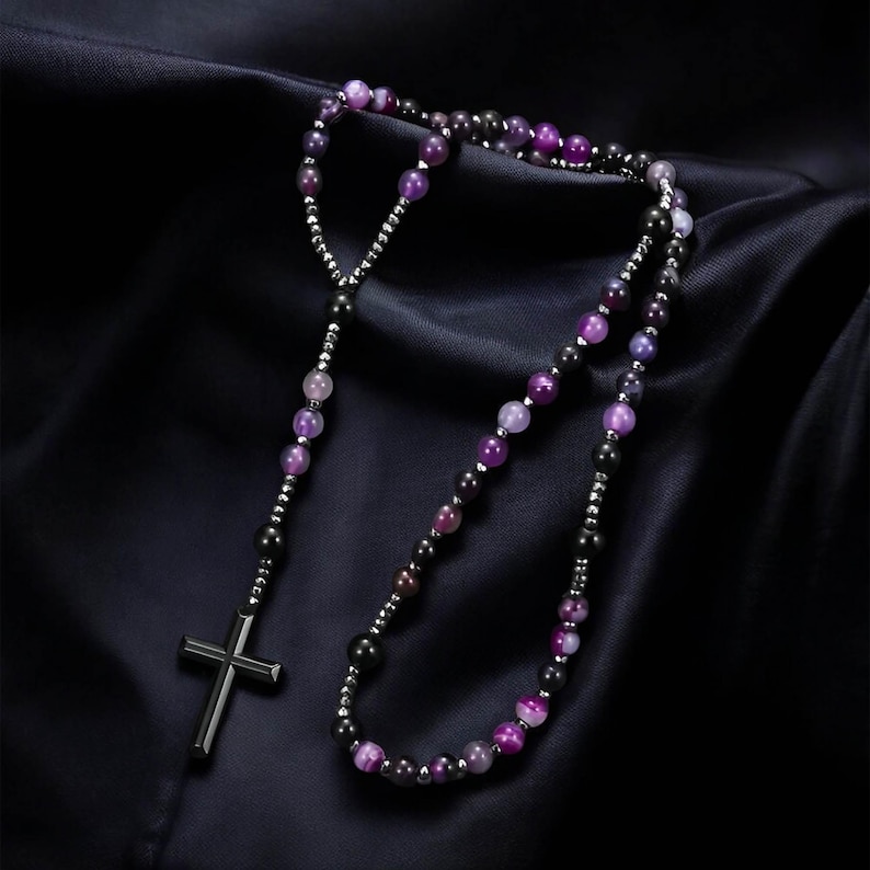 May include: A rosary necklace with a dark metal cross pendant and a strand of purple and black beads. The beads are interspersed with small silver-coloured beads. The necklace is displayed on a dark, textured background.