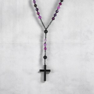 May include: A rosary necklace with a black cross pendant and purple and black beads. The beads are linked by a dark chain. The necklace is set against a white backdrop.