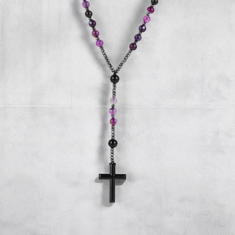 May include: A rosary necklace with a black cross pendant and purple and black beads. The beads are linked by a dark chain. The necklace is set against a white backdrop.