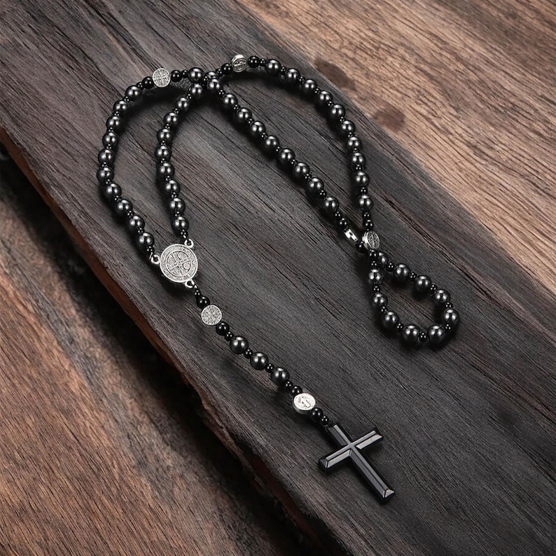 Puede incluir: Un rosario de cuentas negras con un colgante de cruz plateada. El collar presenta cuentas oscuras y redondas y detalles plateados, incluyendo medallones decorativos. El rosario se muestra sobre una superficie de madera oscura.