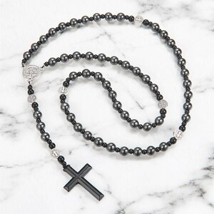 Puede incluir: Un rosario negro con cuentas grises oscuras y un colgante de cruz negro. El collar presenta elementos decorativos plateados y se exhibe sobre una superficie de m&aacute;rmol blanco. Las cuentas est&aacute;n espaciadas uniformemente.