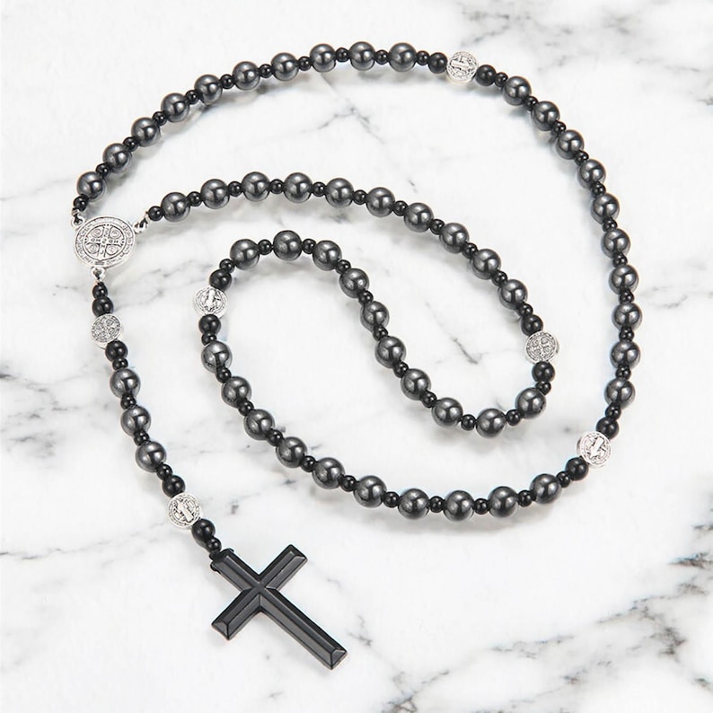 Puede incluir: Un rosario negro con cuentas grises oscuras y un colgante de cruz negro. El collar presenta elementos decorativos plateados y se exhibe sobre una superficie de m&aacute;rmol blanco. Las cuentas est&aacute;n espaciadas uniformemente.