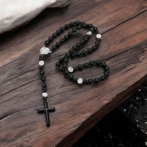 Puede incluir: Rosario de cuentas negras con un colgante de cruz negra y detalles plateados. El rosario presenta una serie de cuentas redondas negras conectadas por una cadena, con una cruz en la parte inferior y medallones religiosos.
