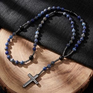 Puede incluir: Un collar de cuentas con un colgante de cruz plateada. El collar presenta cuentas de piedra azul y gris, intercaladas con cuentas negras. La cruz es un diseño metálico simple. El collar descansa sobre una superficie de madera.