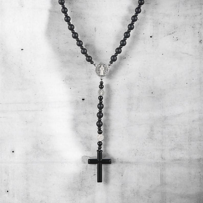 Puede incluir: Collar de rosario negro con cuentas mate y un centro y colgante de cruz plateados. El collar presenta una serie de cuentas redondas conectadas por una cadena, con una cruz en la parte inferior. El centro tiene un dise&ntilde;o detallado.