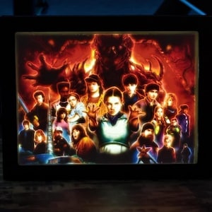 May include: A framed, illuminated artwork featuring a group of characters against a fiery background. The artwork has a black frame and is lit from within, creating a vibrant display. The image is likely a promotional piece or fan art.