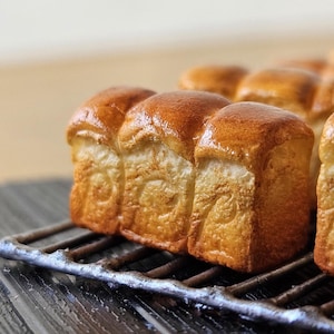 Puede incluir: Primer plano de panecillos cuadrados de color marrón dorado sobre una rejilla metálica. Los panecillos tienen una parte superior brillante y ligeramente caramelizada. La rejilla está sobre una superficie de madera oscura, con un fondo borroso.