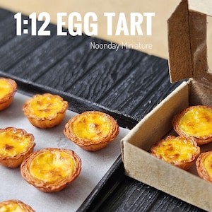 Puede incluir: Miniaturas de tartaletas de huevo con relleno de crema dorada y corteza crujiente, presentadas en una bandeja negra y en una pequeña caja de cartón. Incluye el texto "1:12 EGG TART" y "Noonday Miniature".