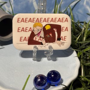 May include: A rectangular pin featuring a cartoon character with blonde hair in a brown military-style uniform, leaning forward with a wide, laughing mouth. The background of the pin displays the repeated red text EA EAEAEAEAEAEAEAEAEAEAEAE.