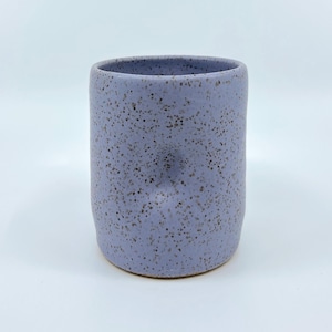 Puede incluir: Taza cilíndrica de cerámica en color lavanda claro con motas. La taza tiene un borde liso y redondeado y una superficie texturizada con motas marrón oscuro. Adecuada para bebidas.