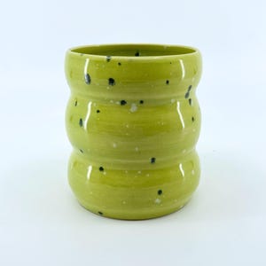 May include: A handmade, lime green ceramic tumbler with a unique, rounded shape. The surface is speckled with black and white dots, adding a touch of visual interest. The tumbler is designed for beverages.