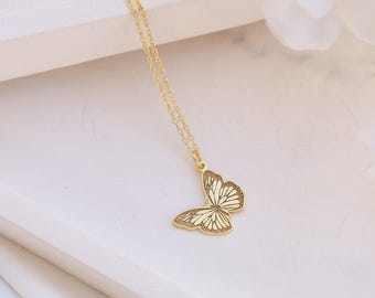 May include: A gold-colored butterfly pendant necklace. The butterfly pendant has a detailed design with cream-colored accents. The necklace is displayed on a white tray with white flowers in the background. The chain is delicate and gold-colored.