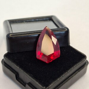 May include: A faceted, teardrop-shaped gemstone with a vibrant red hue, displayed in a black velvet-lined box. The gem's cut reveals multiple facets, catching the light and creating a sparkling effect. The box is open, showcasing the gem's brilliance.