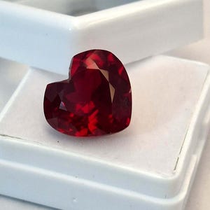 Peut inclure: Une pierre précieuse en forme de cœur, de couleur rouge profond, avec de multiples facettes. Elle est présentée dans un écrin à bijoux blanc. La taille de la pierre crée de multiples surfaces réfléchissantes, rehaussant son éclat.