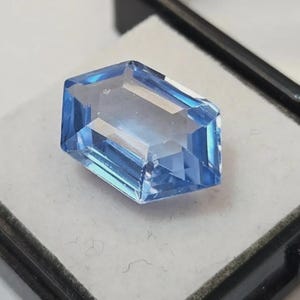 May include: A faceted, light blue gemstone with a rectangular shape and angled edges. The gem is displayed on a white surface within a black display case. The gem's facets reflect light, creating a sparkling effect.