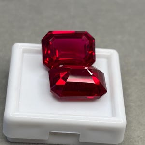 May include: Two vibrant red, emerald-cut gemstones with faceted surfaces that reflect light. The stones are positioned on a white, square display case with a raised border, showcasing their deep crimson color and precise geometric cut.