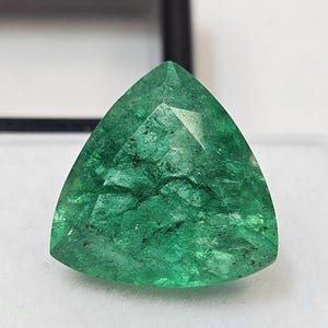以下が含まれることがあります： 三角形の、ファセット加工されたエメラルドの宝石。この宝石は、内部にインクルージョンを持つ鮮やかな緑色をしています。エメラルドは白い背景に置かれ、その透明度と輝きを際立たせています。この宝石は、天然の未処理エメラルドです。
