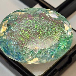May include: A large, oval-shaped gemstone with a faceted surface. The gem displays a mesmerizing play of colors, including shades of green, blue, and hints of purple. It is set in a black display case, showcasing its iridescent qualities.