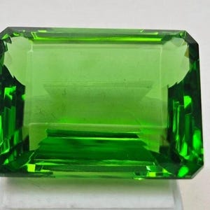 Puede incluir: Una vibrante gema verde de talla esmeralda con forma rectangular. La gema tiene una superficie facetada que refleja la luz, creando un efecto brillante. La gema se muestra sobre una superficie blanca.