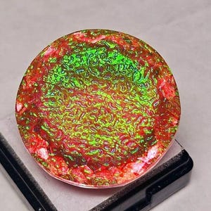May include: A round gemstone with a vibrant, textured surface. The gem features a mix of red, green, and brown hues, creating a unique, eye-catching appearance. The gemstone is set on a black base, highlighting its colors.