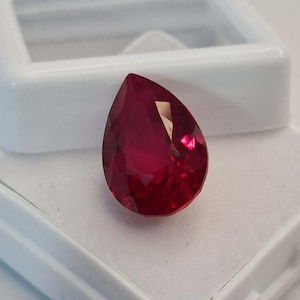 Könnte beinhalten: Ein facettierter, birnenförmiger Rubin-Edelstein. Der Edelstein hat eine tiefe, leuchtende rote Farbe und ist vor einem weißen Hintergrund platziert. Der Edelstein wird in einem weißen Kunststoffetui präsentiert.