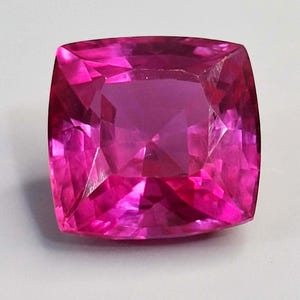 Puede incluir: Una gema facetada, de talla cojín, en un vibrante tono rosa. Las facetas de la gema reflejan la luz, creando un efecto brillante. La forma cuadrada con esquinas redondeadas es un diseño clásico para joyería.