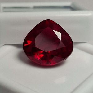 Peut inclure: Une pierre précieuse facettée d'un rouge profond avec une forme triangulaire. La pierre présente de multiples coupes géométriques qui reflètent la lumière, créant un aspect brillant. Elle est positionnée au centre, affichant une couleur cramoisie riche.