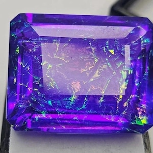 May include: A large, rectangular gemstone with a deep purple hue and iridescent flashes of blue, green, and yellow. The gem has a faceted cut, and the light reflects off the internal structures. The gemstone is a striking piece of jewelry.