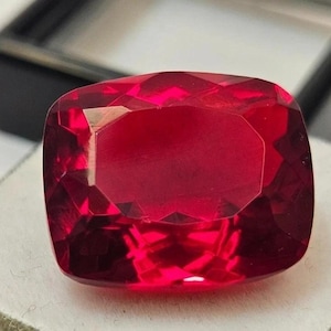 May include: A faceted, cushion-cut ruby gemstone. The gem is a deep, vibrant red colour with multiple facets that reflect light. The ruby is set against a neutral background, highlighting its colour and clarity. This gemstone is likely for jewellery.