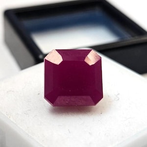 May include: A faceted, deep red ruby gemstone with an emerald cut. The gemstone is displayed on a white surface, with a black box in the background. The ruby's facets reflect light, showcasing its rich color and clarity. Jewelry and gemstone.