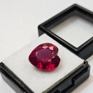 May include: A heart-shaped, deep red gemstone with multiple facets sits on a white surface inside a black display case. The gemstone's cut creates many reflective surfaces, enhancing its brilliance. The case is open, revealing the gem's vibrant colour.