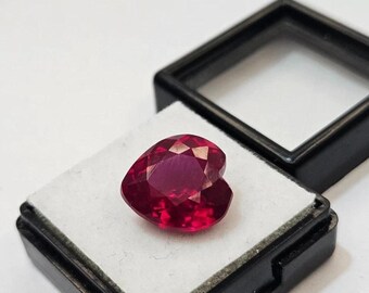Puede incluir: Una gema roja oscura facetada en forma de corazón descansa sobre una superficie blanca dentro de una vitrina negra. El corte de la gema crea múltiples superficies reflectantes, realzando su brillo. La vitrina está abierta.