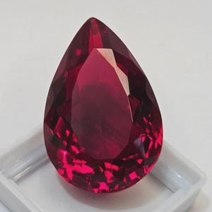 Könnte beinhalten: Ein facettierter, birnenförmiger Edelstein in tiefroter Farbe. Der Edelstein befindet sich in einer weißen Kunststoffschale, die seine Klarheit und Brillanz hervorhebt. Der Schliff des Steins erzeugt mehrere reflektierende Oberflächen.