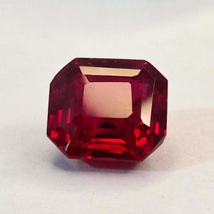 Könnte beinhalten: Ein facettierter, rechteckig geschliffener, tiefroter Edelstein. Der Edelstein hat eine helle, reflektierende Oberfläche mit mehreren Facetten, die das Licht einfangen. Der Edelstein liegt vor einem schlichten weißen Hintergrund.