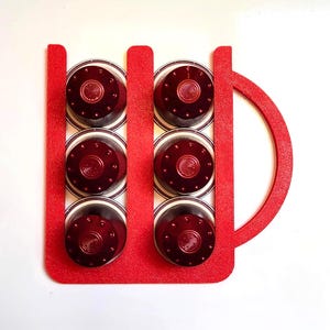 May include: A red coffee pod holder in the shape of a mug, containing six dark red coffee pods. The pods are arranged in two columns of three, with a clear top and a patterned design. The holder is made of a solid red material.