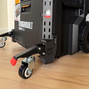 Puede incluir: Una caja de herramientas rodante Toughbuilt Stacktech negra con detalles en naranja. La caja de herramientas tiene dos cajones, un asa y una gran rueda todoterreno. Tiene dos pequeñas ruedas delanteras y un acento rojo en el soporte de la rueda delantera.