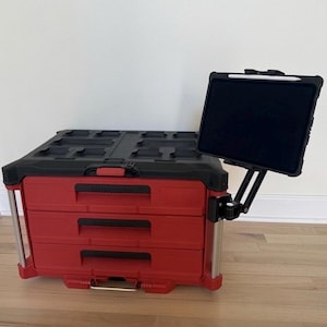 Peut inclure: Une boîte à outils modulaire rouge et noire avec trois tiroirs et une surface supérieure noire. Un bras articulé noir réglable est fixé sur le côté, supportant une tablette noire dans un étui de protection avec un stylet blanc sur le bord.