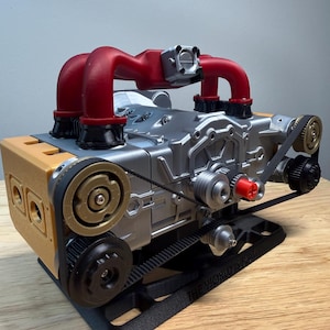 May include: A detailed model engine with a silver body, red intake manifolds, and gold and black accents. The model sits on a black stand and features belts and pulleys. The text "THE WORLD IS FLAT" is visible on the stand.