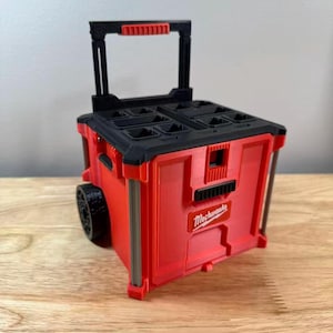 May include: A red and black rolling tool box with a telescoping handle and wheels. The front features a rectangular red logo plate with the white text "Milwaukee" printed on it. The top lid has multiple recessed compartments and a black handle.