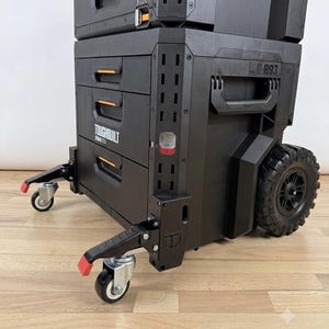 Può includere: Un'unità mobile nera ToughBuilt StackTech per riporre attrezzi, dotata di tre cassetti, dettagli arancioni e la scritta R93. L'unità presenta due grandi ruote robuste sul retro e due ruote piroettanti bloccabili più piccole sul davanti.