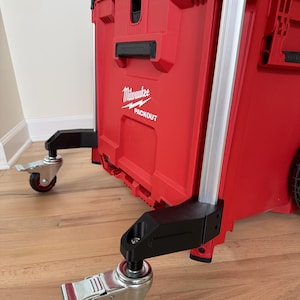 Könnte beinhalten: Eine rote Milwaukee Packout Roll-Werkzeugbox mit schwarzem Deckel und schwarzem Griff. Das Gerät ist mit vier Lenkrollen mit Feststellmechanismus ausgestattet, die an schwarzen Halterungen an den Ecken der Unterseite befestigt sind.