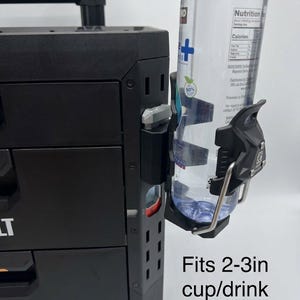 Puede incluir: Un portavasos de plástico negro montado en el lateral de una unidad de almacenamiento de herramientas negra. El soporte cuenta con un marco de alambre metálico y sostiene una botella de agua de plástico transparente. El texto dice: Fits 2-3in cup/drink.