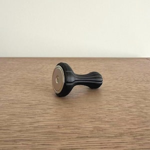 May include: A black coffee tamper with a contoured handle and a flat, circular metal base. The handle features vertical ridges along its length, and the base is secured with a central screw, showing a metallic finish on the bottom surface.