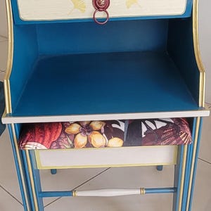 Peut inclure: Une table de chevet peinte à la main avec un intérieur bleu et des bordures dorées. Le tiroir est de couleur crème avec des accents de feuilles jaunes et une poignée rouge. Un coussin à motifs repose sur l'étagère inférieure.