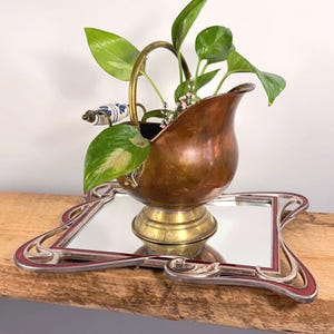 May include: A copper-coloured decorative container with a brass base and handle, holding green leafy plants. It sits on a mirrored tray with a silver frame and red accents. The handle has a white and blue patterned ceramic detail.