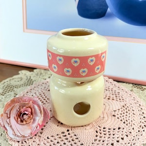 May include: A cream-coloured ceramic container in two parts, with a pink band adorned with blue hearts. A paper rose and a pink doily are in the foreground. A framed picture with blue vases is in the background.