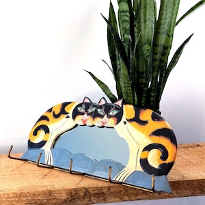 May include: A whimsical cat-themed wall mirror with a blue, wavy base and three hooks. The mirror features two calico cats with orange, black, and white markings. A tall green plant is placed behind the mirror on a wooden shelf.