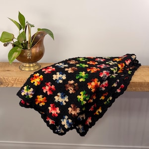 May include: A black crocheted blanket featuring colourful flower patterns in red, yellow, blue, and green. The blanket is draped over a wooden shelf. A brass planter with a green plant is to the left of the blanket.