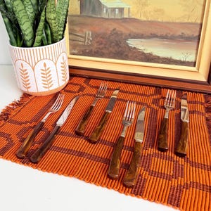 May include: A set of vintage cutlery, including forks and knives, with dark brown handles and silver blades. The utensils are arranged on an orange and brown woven placemat. A framed landscape painting and a potted plant are in the background.