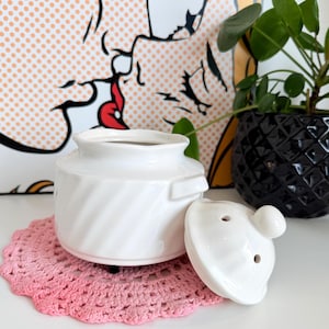 May include: A white ceramic jar with a lid, resting on a pink crocheted doily. The jar has a spiral design and a small handle. The lid has a small knob and two holes. A Pop Art print is in the background.